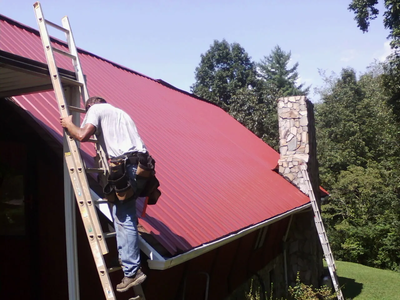Roofer on a ladder beside a red metal roof and stone chimney — Soffit & Fascia Repair by Roofing Brothers in Upsala